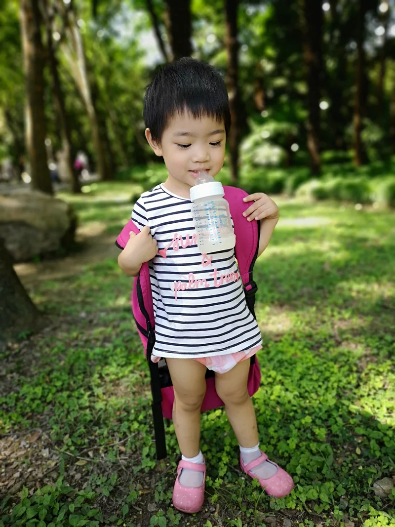2018年，妹妹穿着条纹上衣和尿不湿，背着玫红色书包，嘴上叼着奶瓶，站在树荫下
