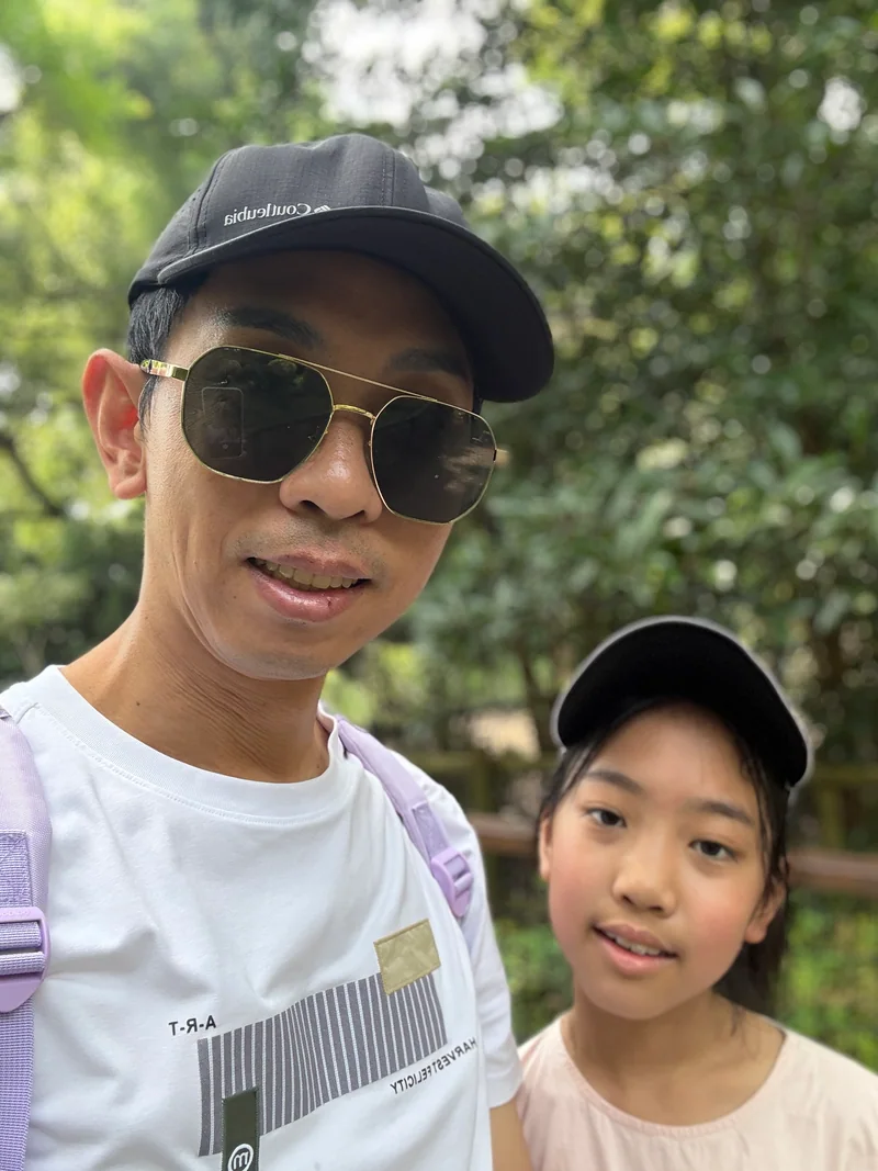 2026年,爸爸与妹妹戴着帽子,在动物园的树荫下合影