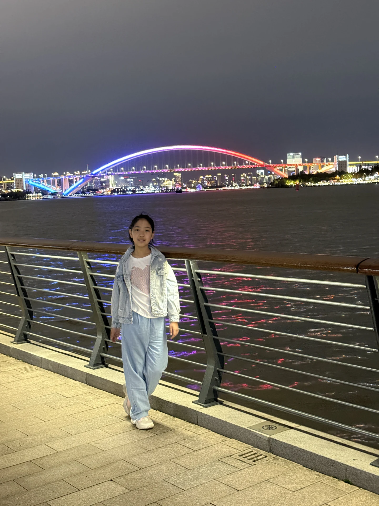 妹妹站在江边栏杆旁,身后是粉紫色灯光的大桥