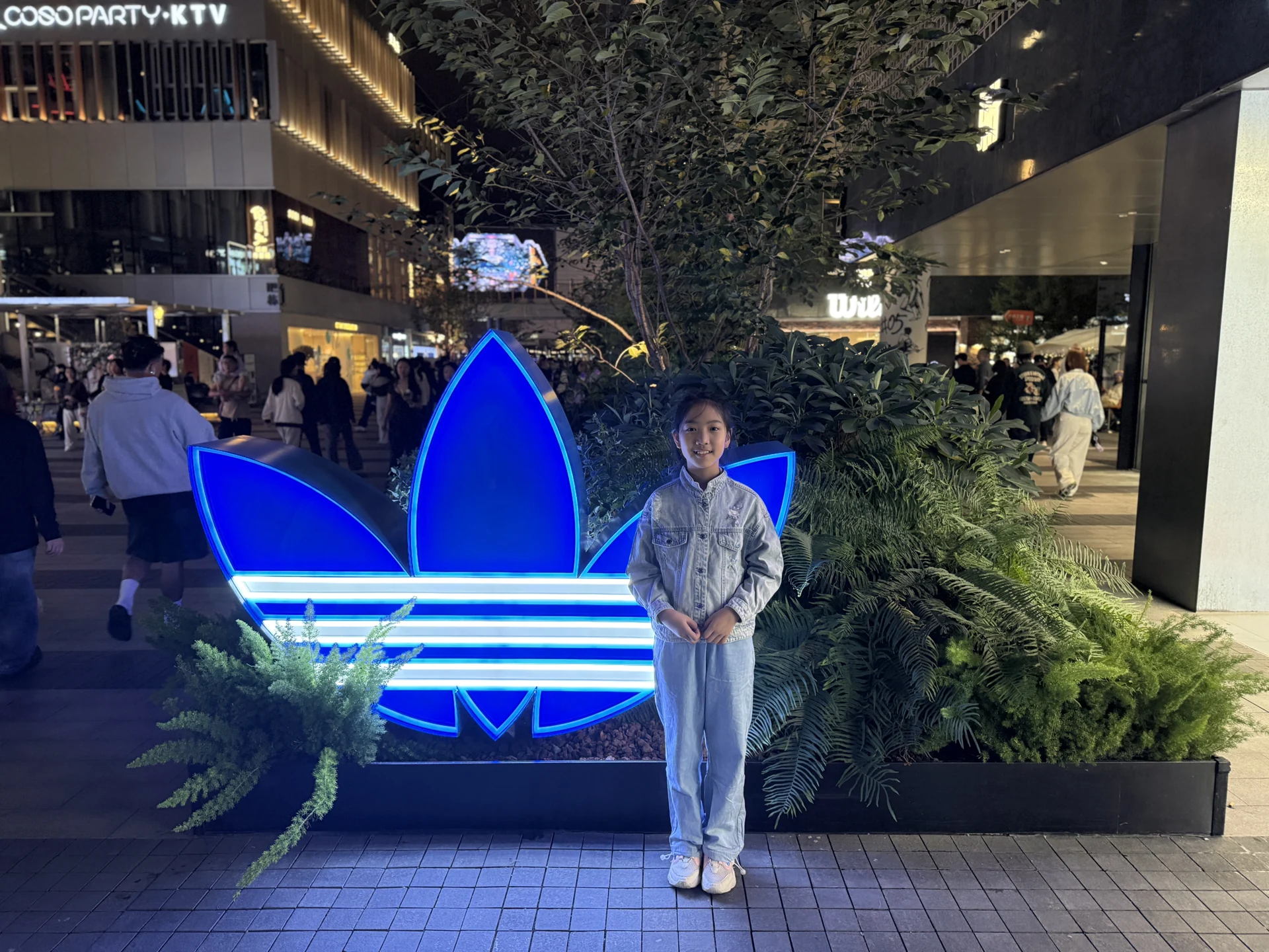 妹妹睁着眼睛,站在发出蓝色灯光的 adidas 三叶草装置旁,身后是街区和人群