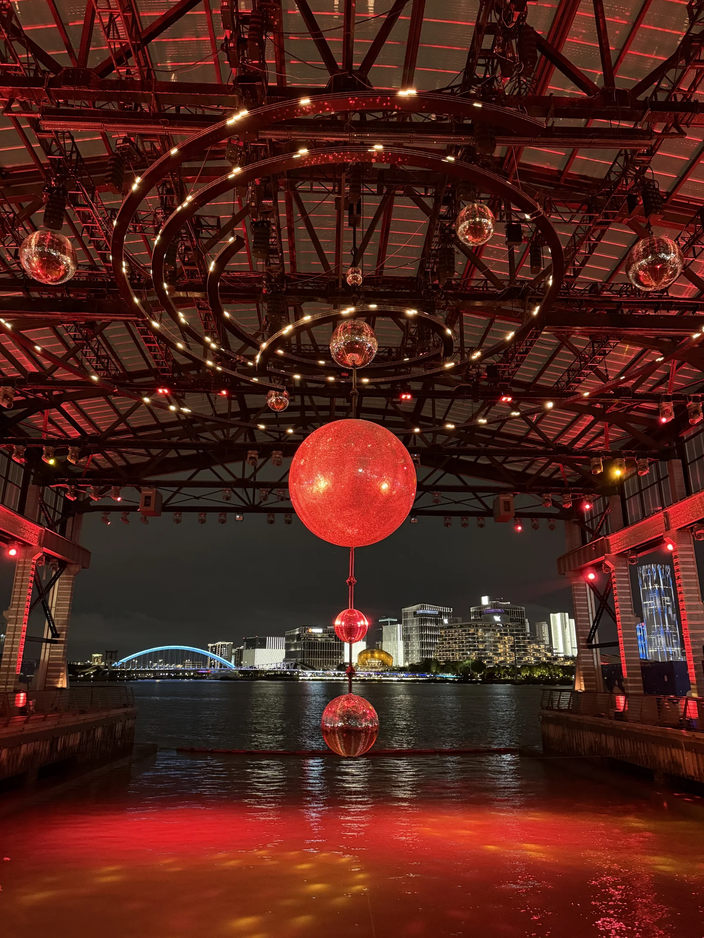 钢架下悬挂着发光的球,红色灯光铺在地面,外面是黄浦江夜景