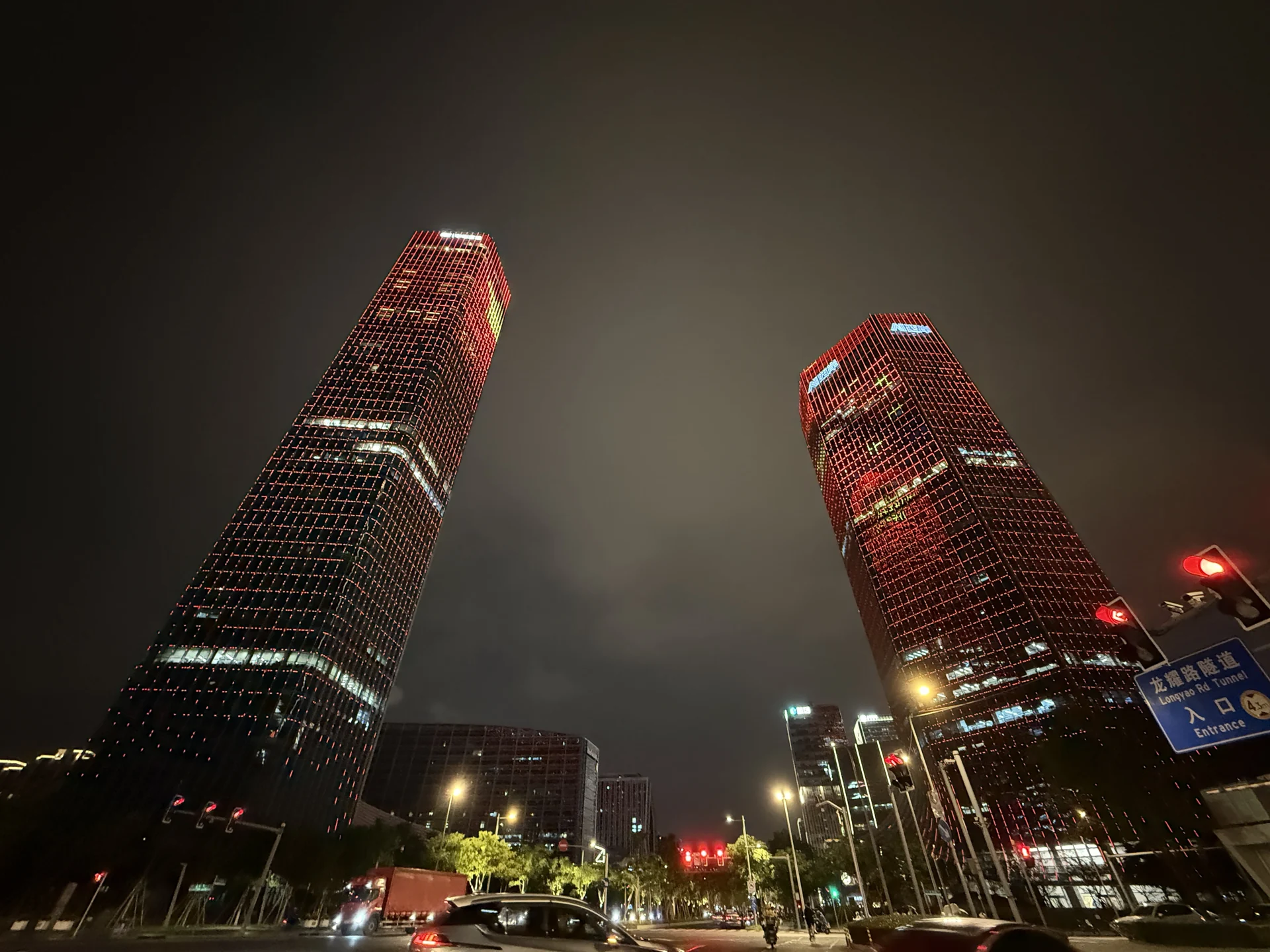 从路边仰望亮着红色灯光的两座高楼,下方是车辆与街道